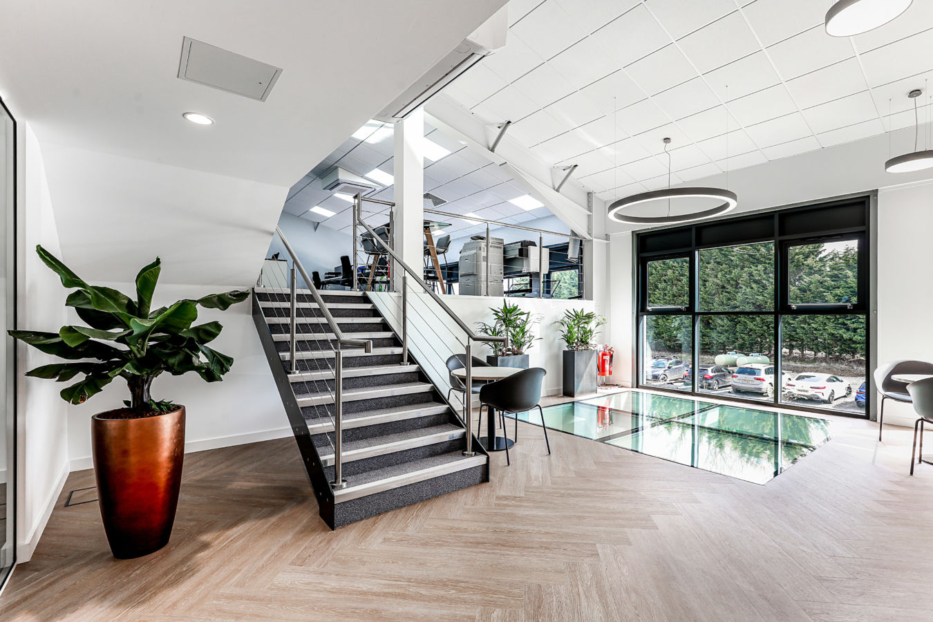 A modern office interior features a staircase leading to an open workspace, with plants decorating the area and large windows overlooking parked cars outside.