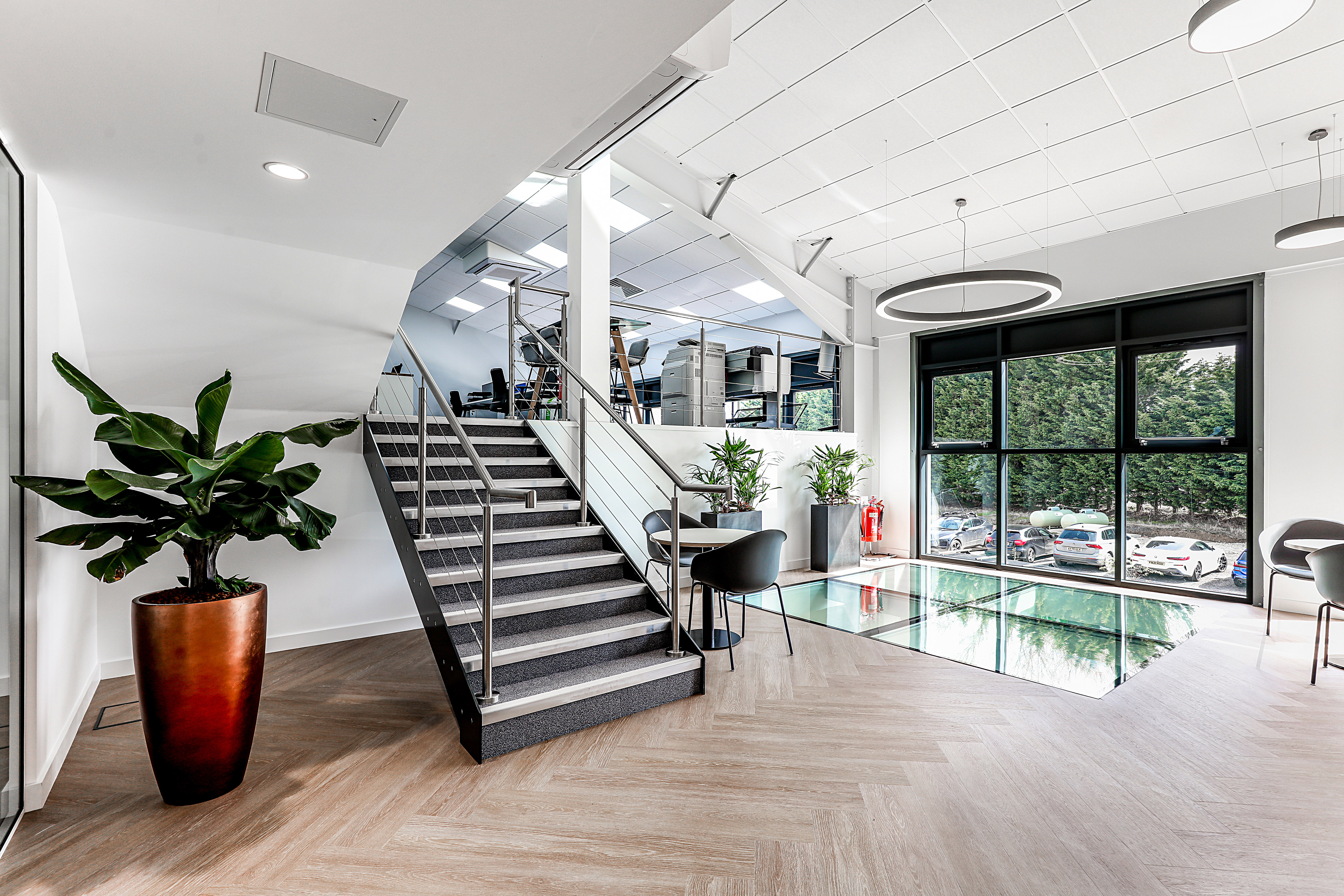 A modern office interior features a staircase leading to an open workspace, with plants decorating the area and large windows overlooking parked cars outside.
