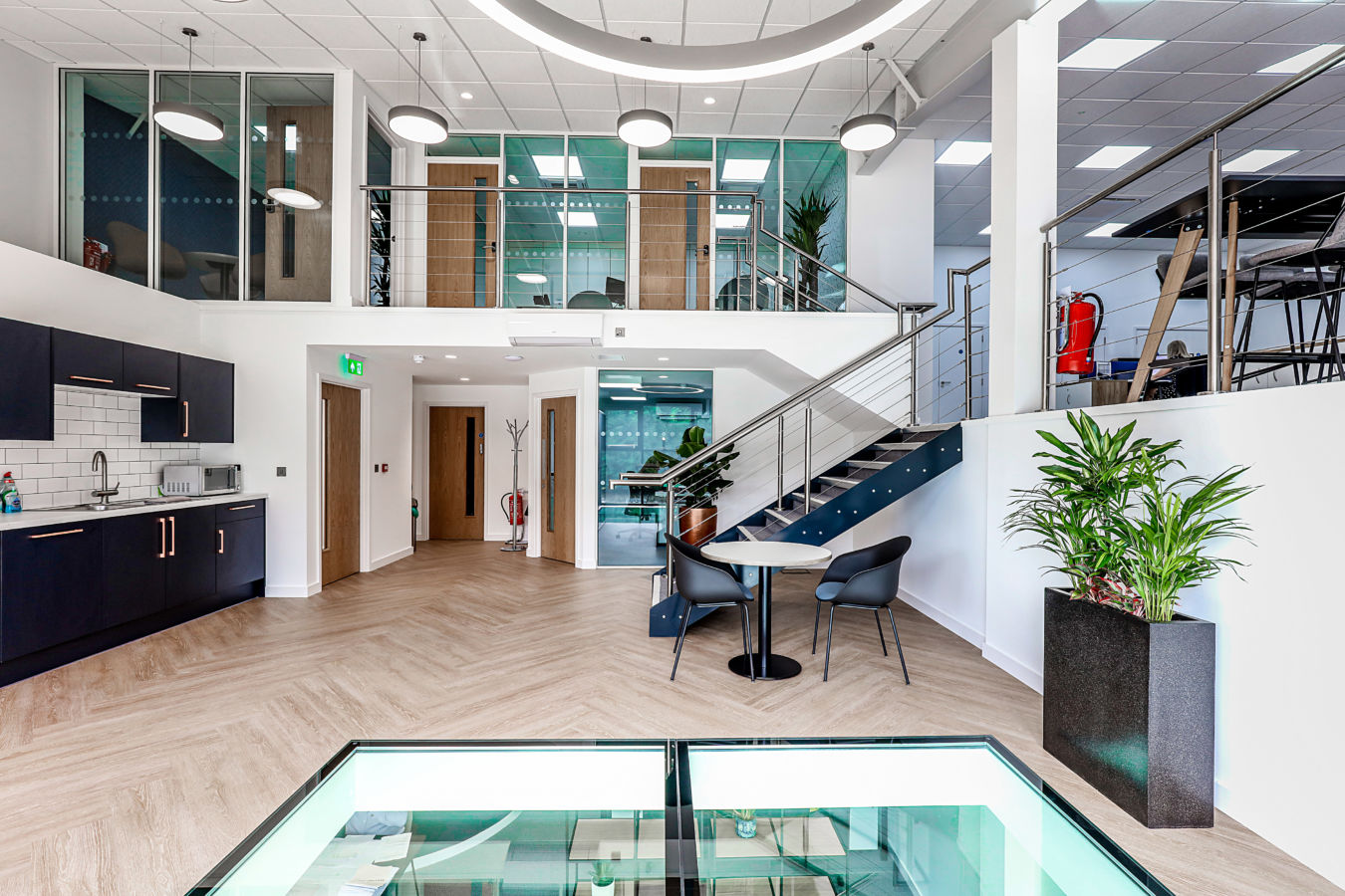 Modern office interior with a kitchenette, dining table, and staircase leading to upper offices, featuring wooden floors, glass railings, potted plants, and a white ceiling with hanging lights.