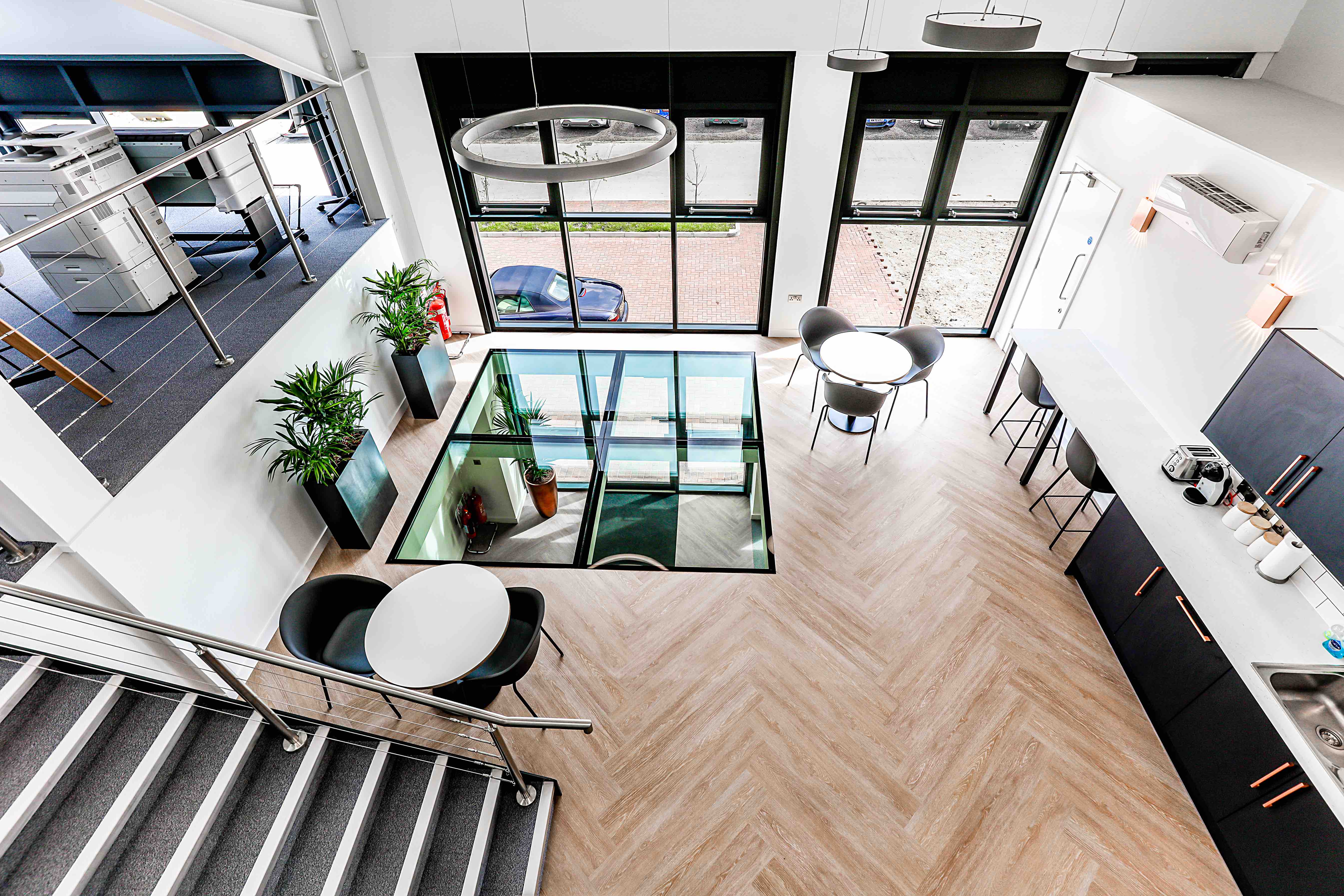 Modern office space, seen from above, with wooden flooring, glass ceiling section, black chairs, round white tables, and large windows, creating a bright, open atmosphere