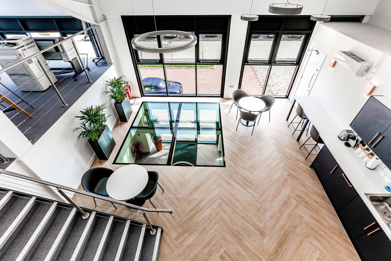 Office interior with a loft-style layout features tables and chairs, a staircase, glass floor section, and kitchen area with appliances, surrounded by large windows and plants.