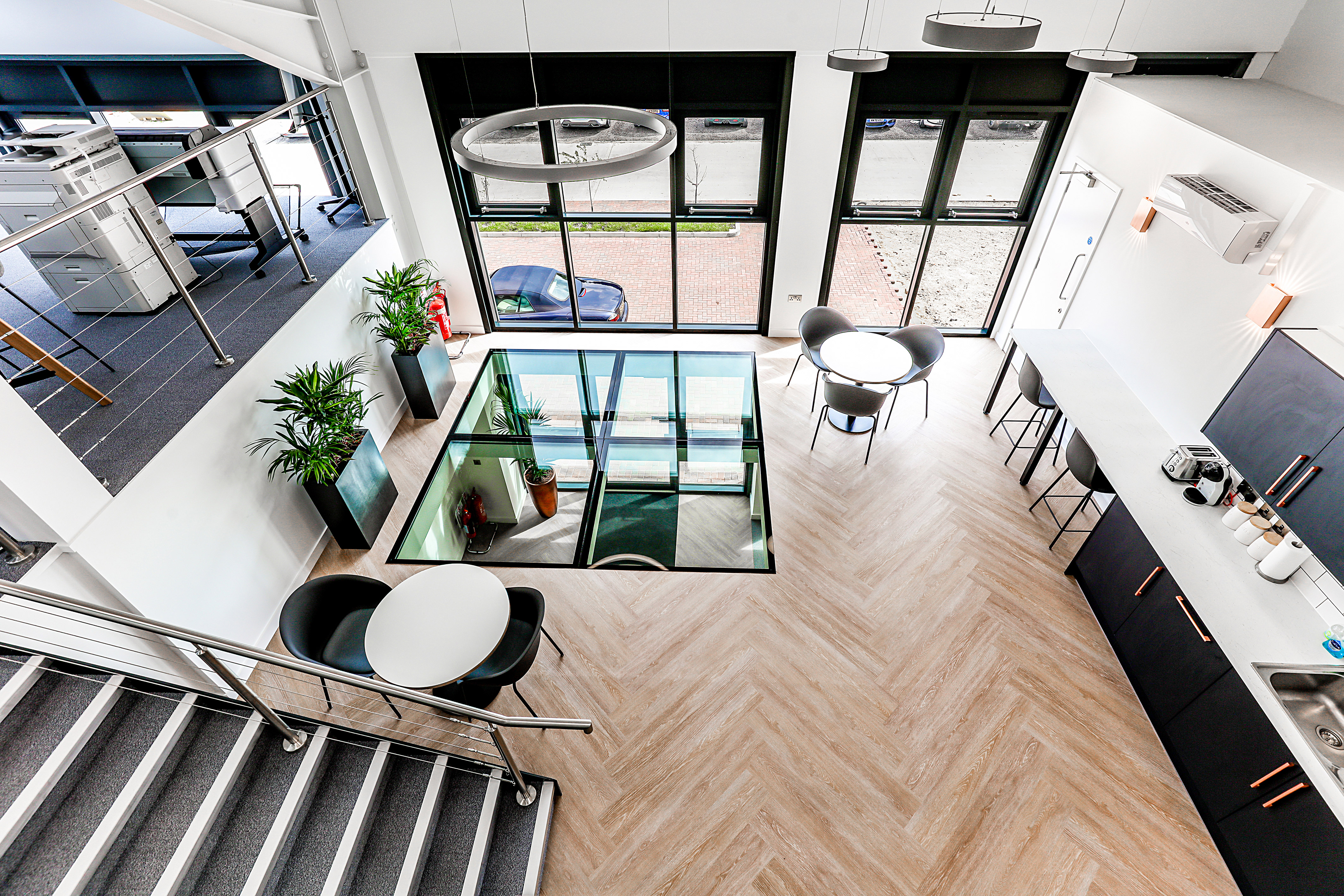 Office interior with a loft-style layout features tables and chairs, a staircase, glass floor section, and kitchen area with appliances, surrounded by large windows and plants.