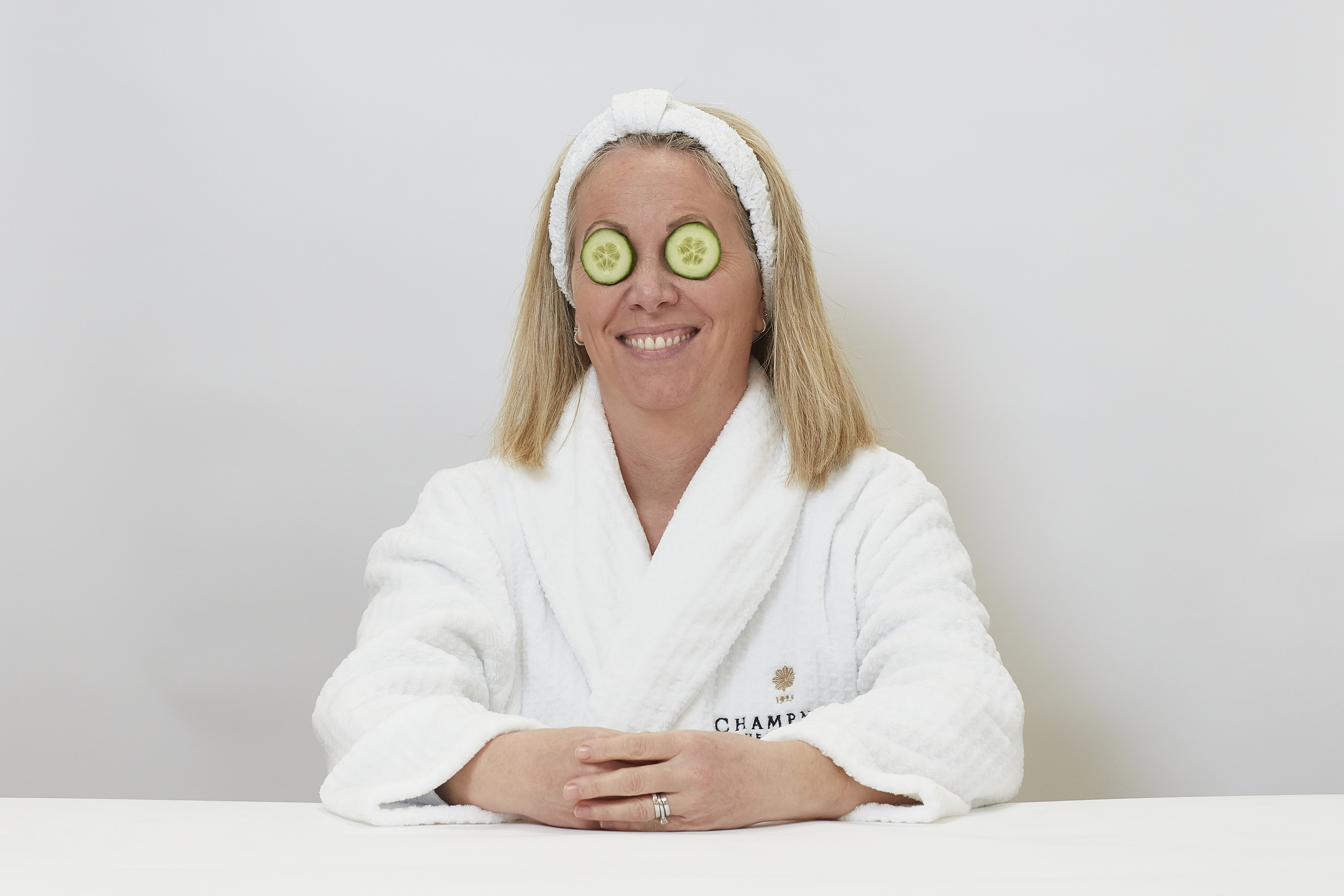 A person wearing a white robe with "CHAMPNEYS" logo and "1925" text, sits smiling with cucumber slices over their eyes, against a plain white background.