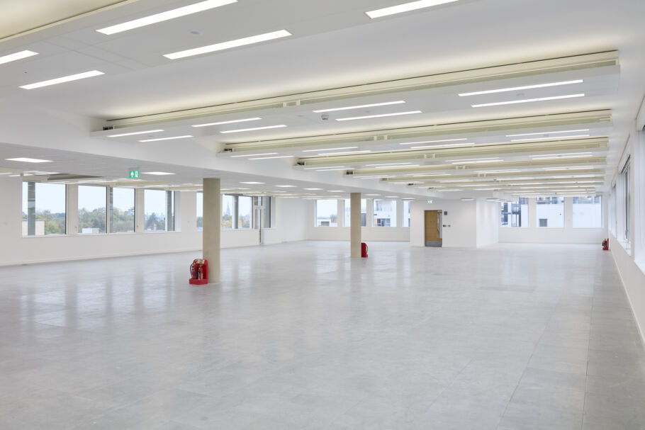 Spacious empty room with gray tiled floor, large windows, and ceiling lights; multiple red fire extinguishers stand against pillars, bordered by white walls and outdoor city view.