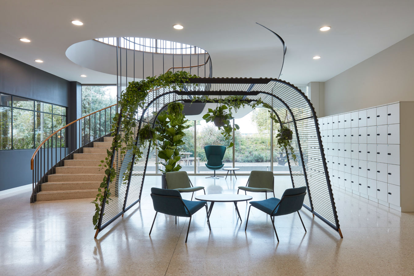 Contemporary office atrium with circular staircase, greenery-covered seating structure, modern chairs, round table, and wall of lockers.