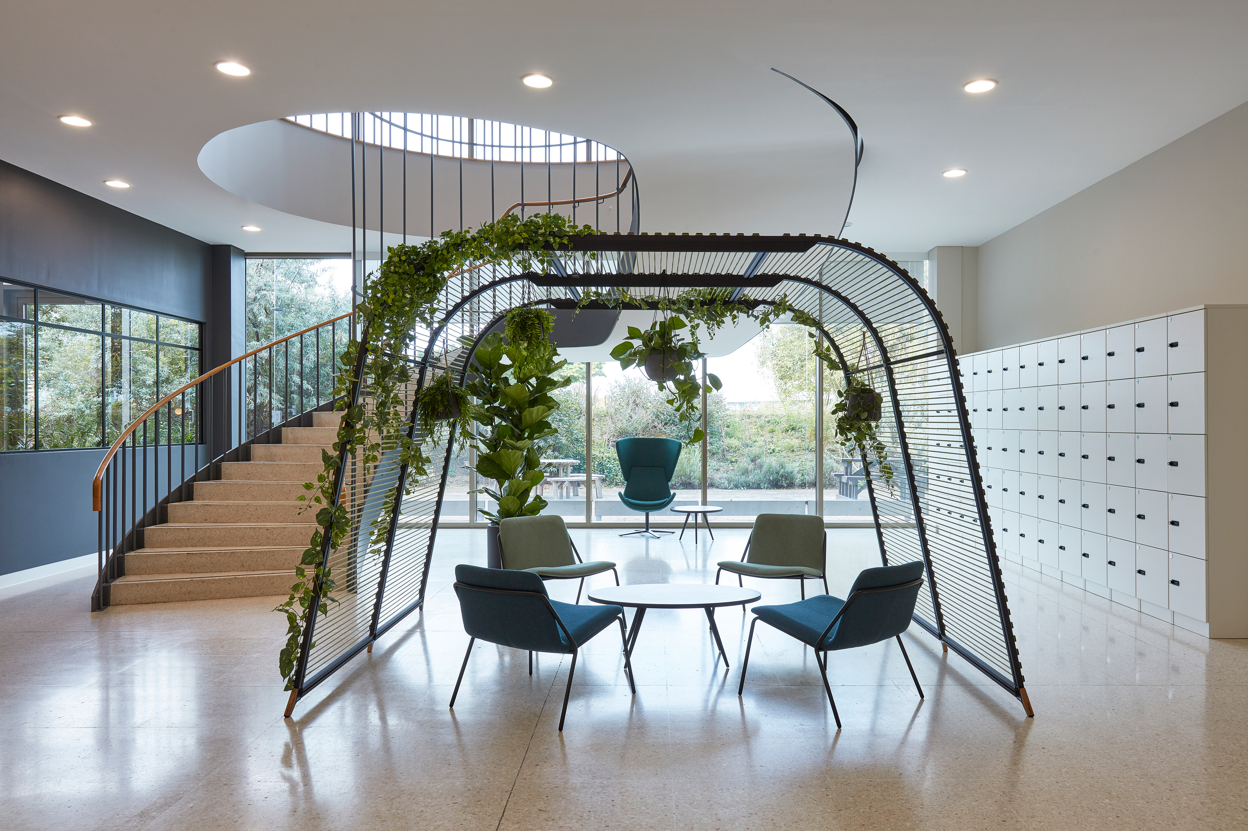 Contemporary office atrium with circular staircase, greenery-covered seating structure, modern chairs, round table, and wall of lockers.
