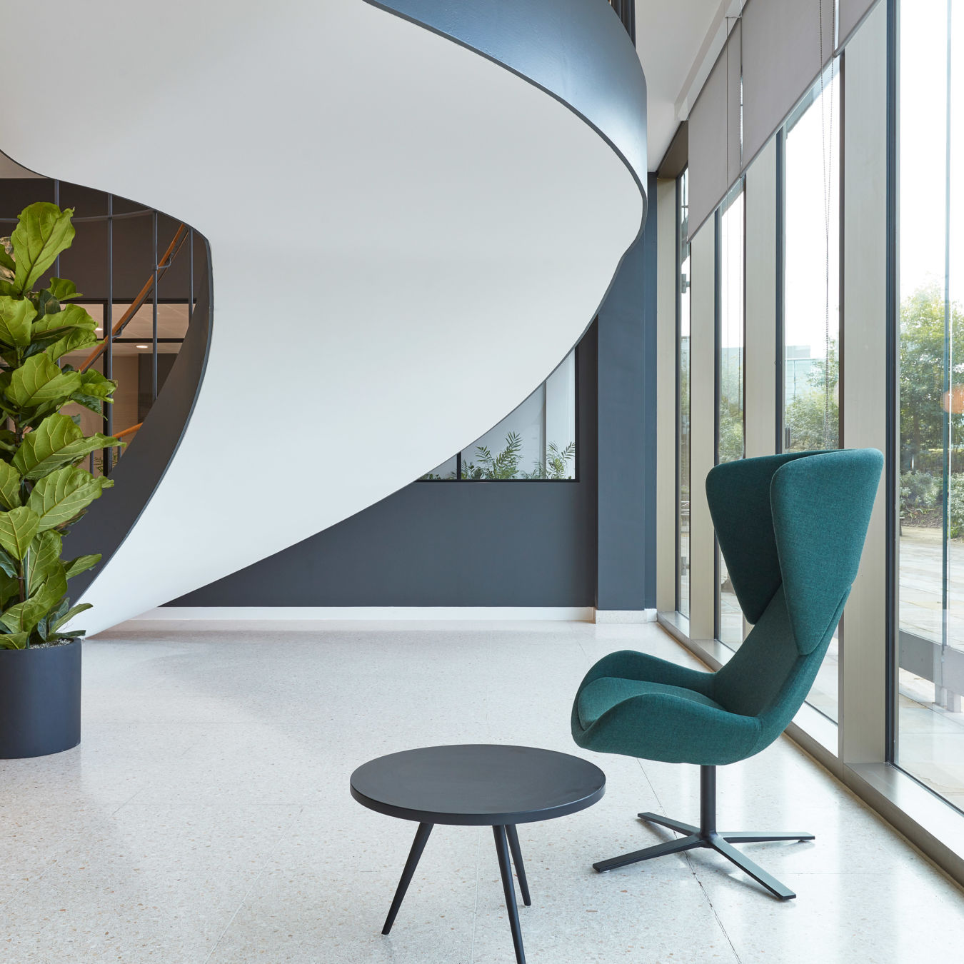 Modern lobby with a sculptural white spiral staircase, green lounge chair, black round table, large windows, and indoor plants.