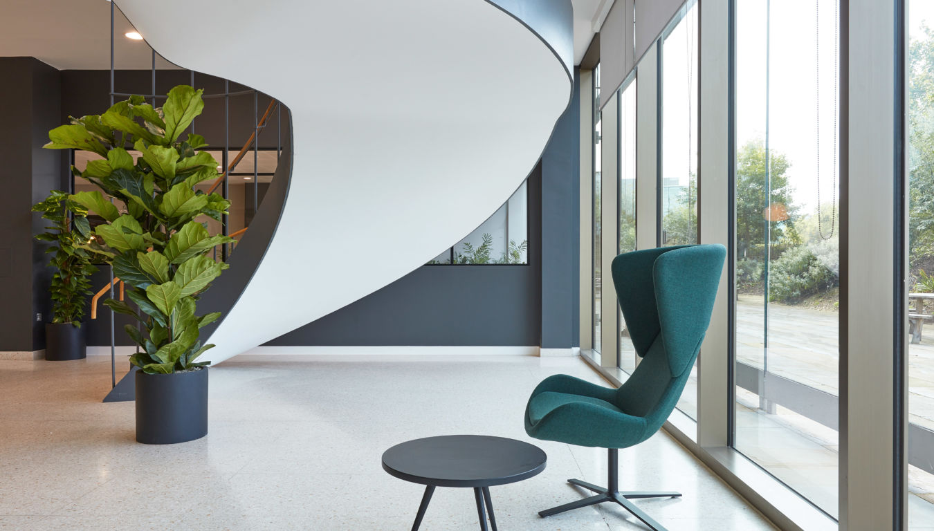 Modern lobby with a sculptural white spiral staircase, green lounge chair, black round table, large windows, and indoor plants.