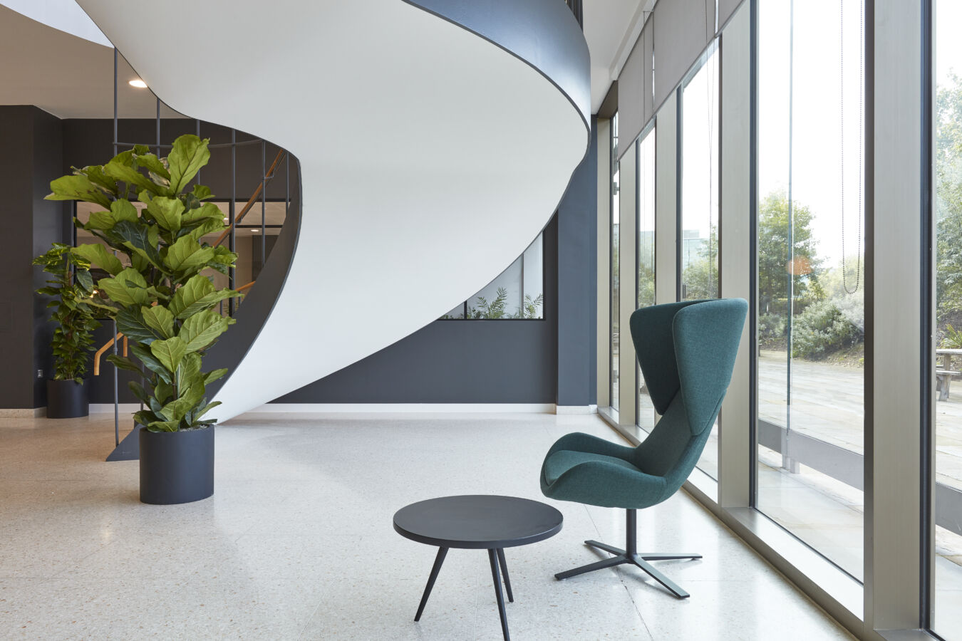 Spiral staircase curves elegantly above a modern lobby, featuring a green upholstered chair and round black table beside tall windows and potted plants.