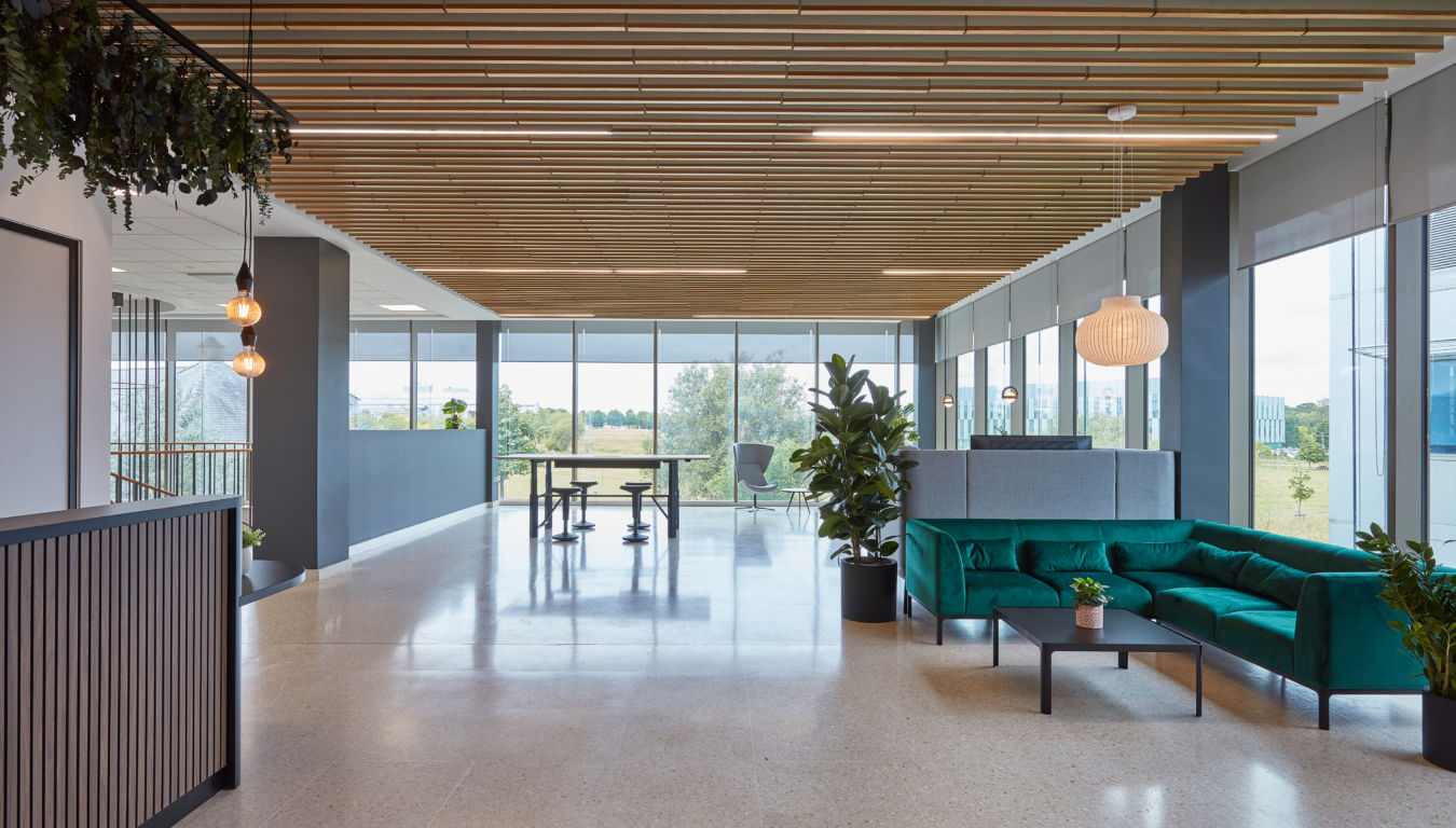 Modern office lounge with a green sofa and potted plants. Large windows offer a view of greenery. Wooden ceiling and pendant lights create a cozy atmosphere