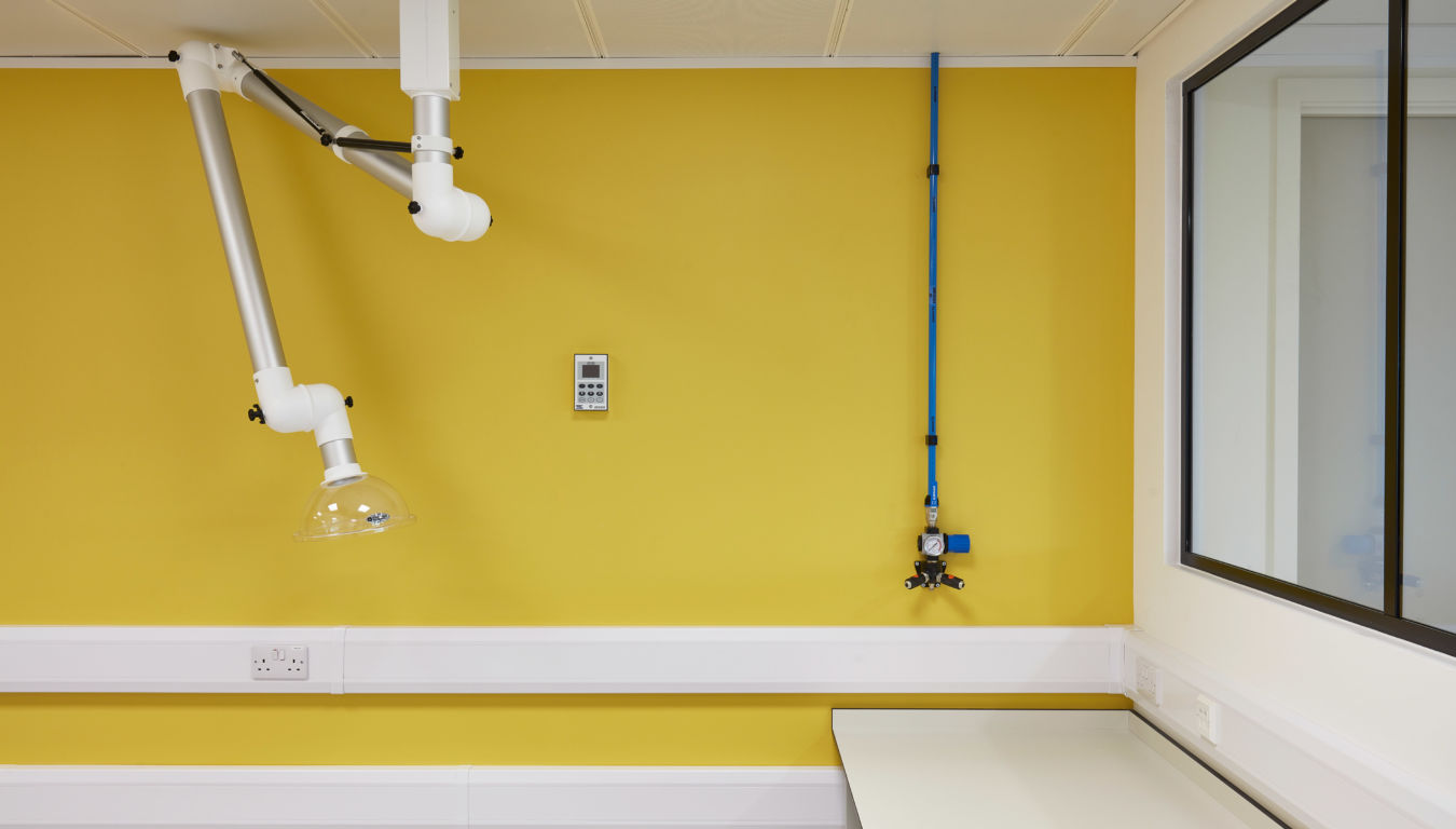 An articulated arm with a transparent dome extends from the ceiling in a yellow-walled laboratory, near a digital control panel and a blue pipe system beside a window.