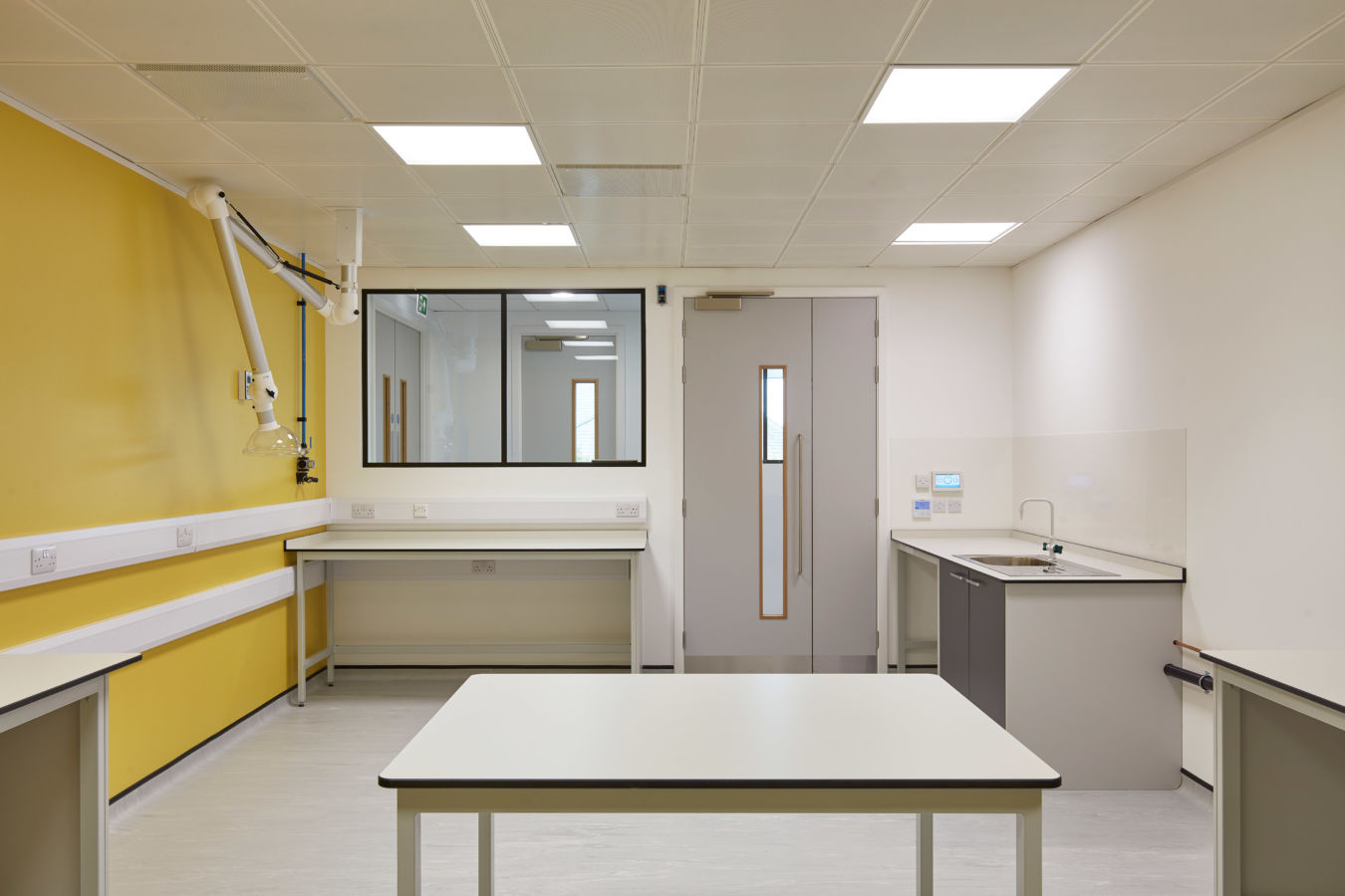 Bright lab room with yellow and white walls, ceiling lights, a central white table, side counters, and a sink. Modern, clean, and organized atmosphere