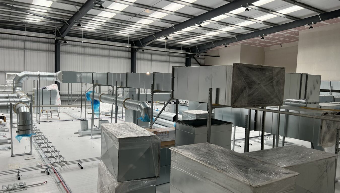 Industrial facility under construction featuring a partially installed HVAC system with exposed ductwork, metal framing, and wrapped ventilation units beneath a steel truss ceiling with skylights.