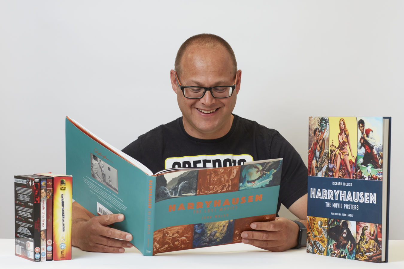 Man smiling while reading a book titled "Harryhausen: The Lost Movies" by John Walsh, beside DVDs and another book, "Harryhausen: The Movie Posters" by Richard Holliss, in a bright setting.