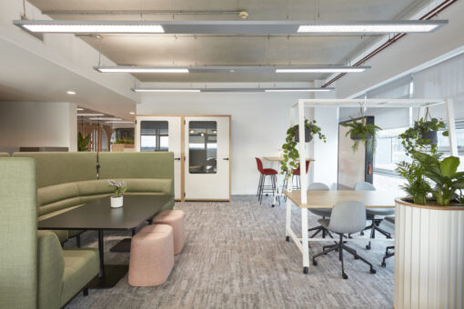 Open-plan office with collaborative seating, green upholstered bench, pink stools, modular desks, and private meeting pods.