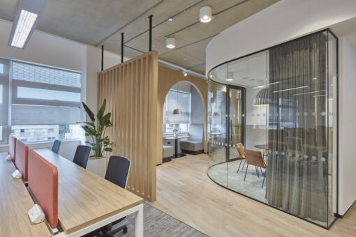 Modern office interior with wooden slat dividers, curved glass meeting room, arched seating nooks, and contemporary furnishings.