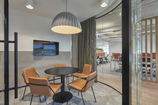 Small meeting room with round table, four tan chairs, large pendant light, and a wall-mounted screen, adjacent to an open office space