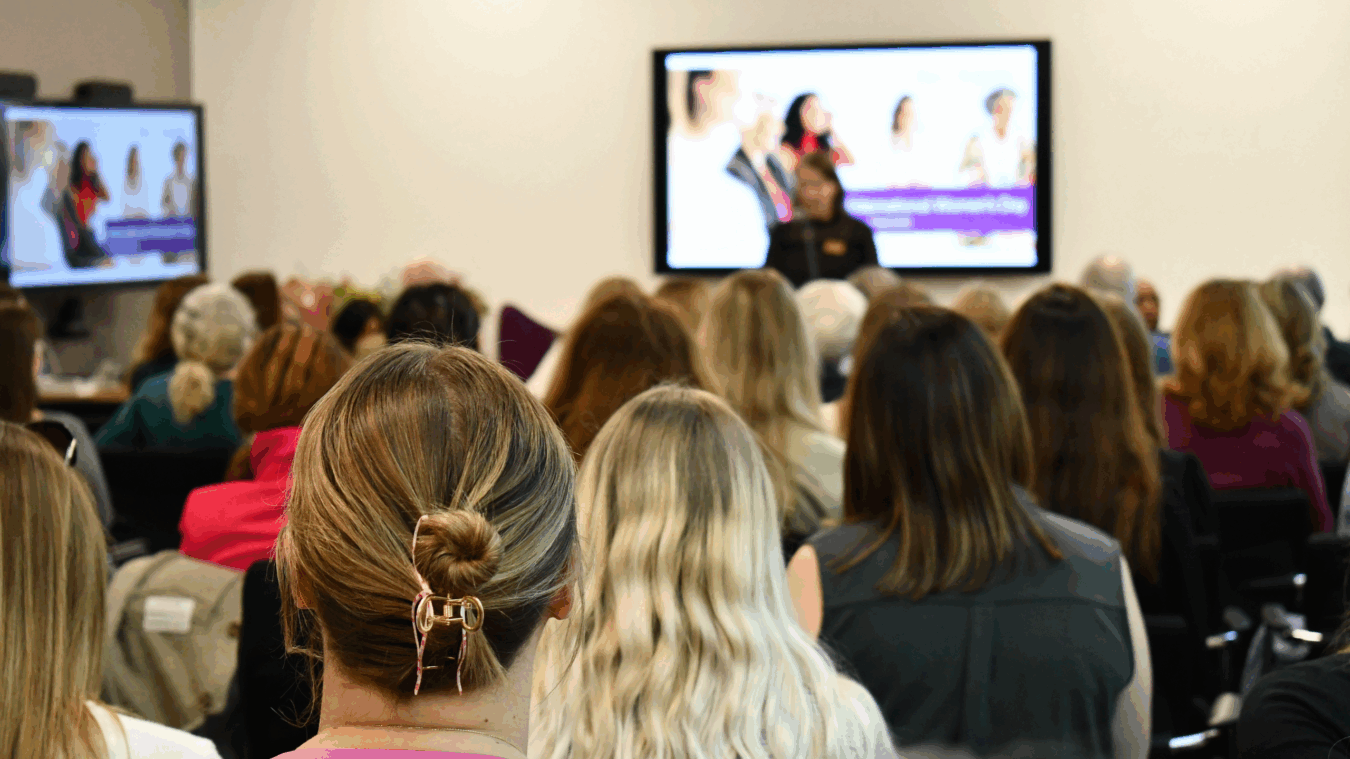 Audience members listen to a presentation with a woman speaking at the front; screens display "International Women's Day March 2023" surrounded by images of people in a conference room.