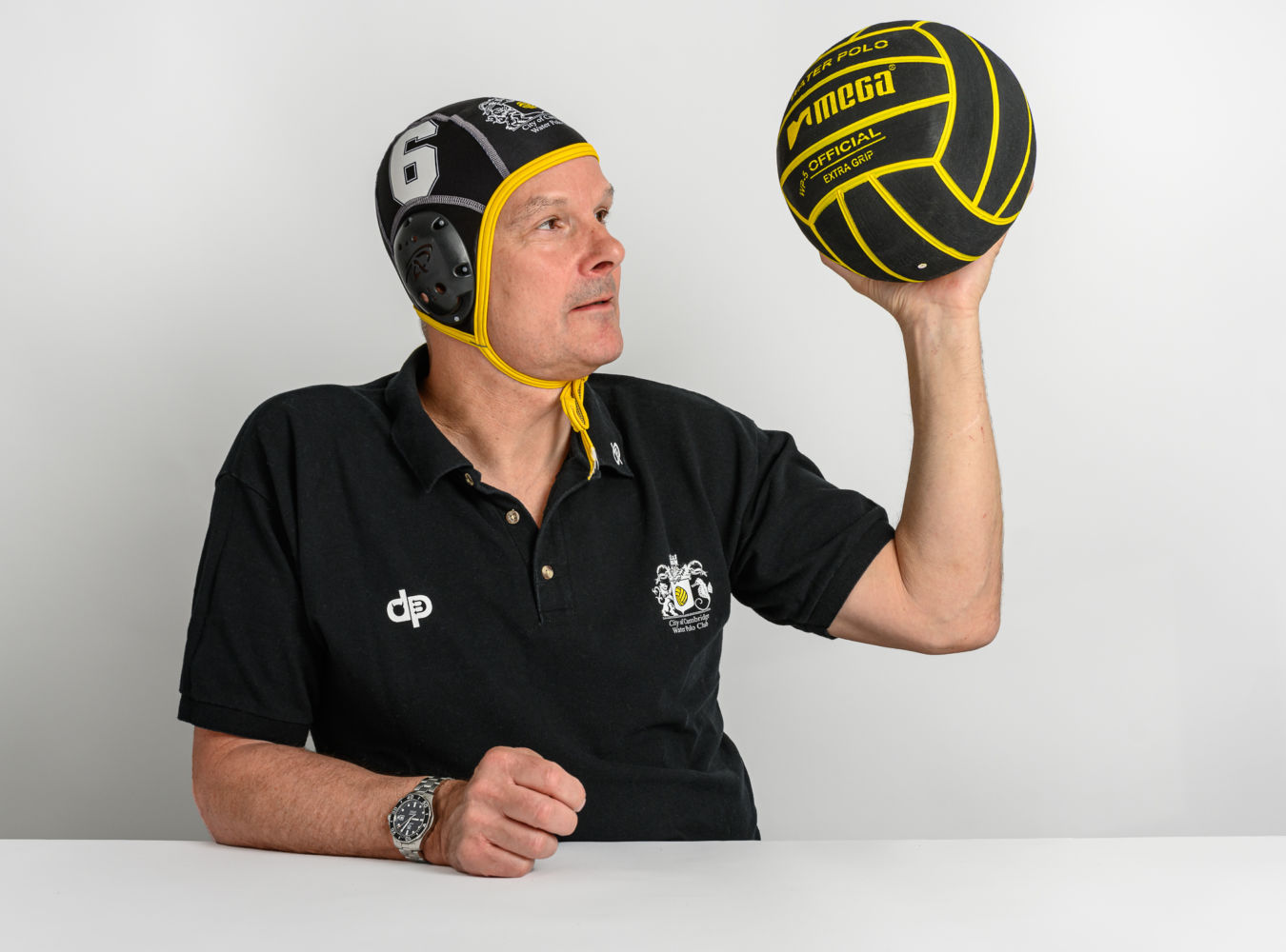 A person wearing a black water polo cap and polo shirt holds a yellow-and-black water polo ball labeled “MEGA OFFICIAL EXTRA-GRIP” against a plain white background.