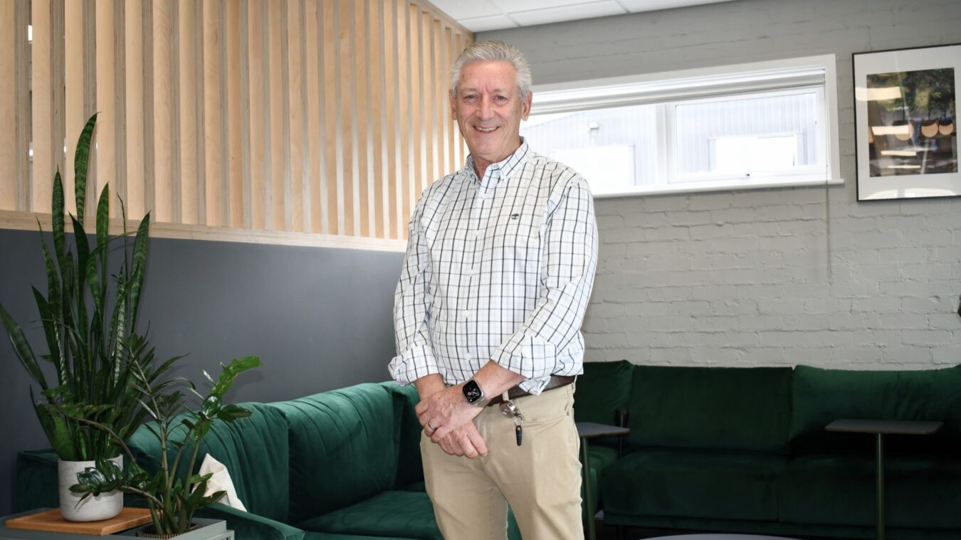 A person stands smiling in a modern office with green sofas, potted plants, and a wooden panel wall. They wear a checked shirt and beige trousers, with a smartwatch on their wrist.