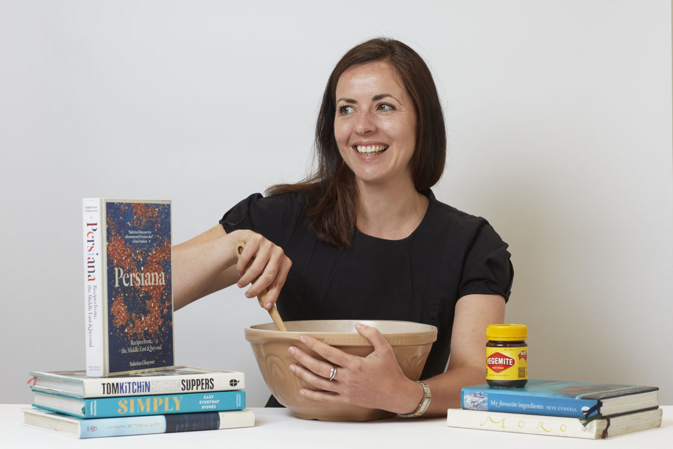A woman stirs a large bowl using a wooden spoon, smiling in a kitchen setting. Cookbooks, such as "Persiana" and "Simply," accompany a jar of Vegemite in the background.