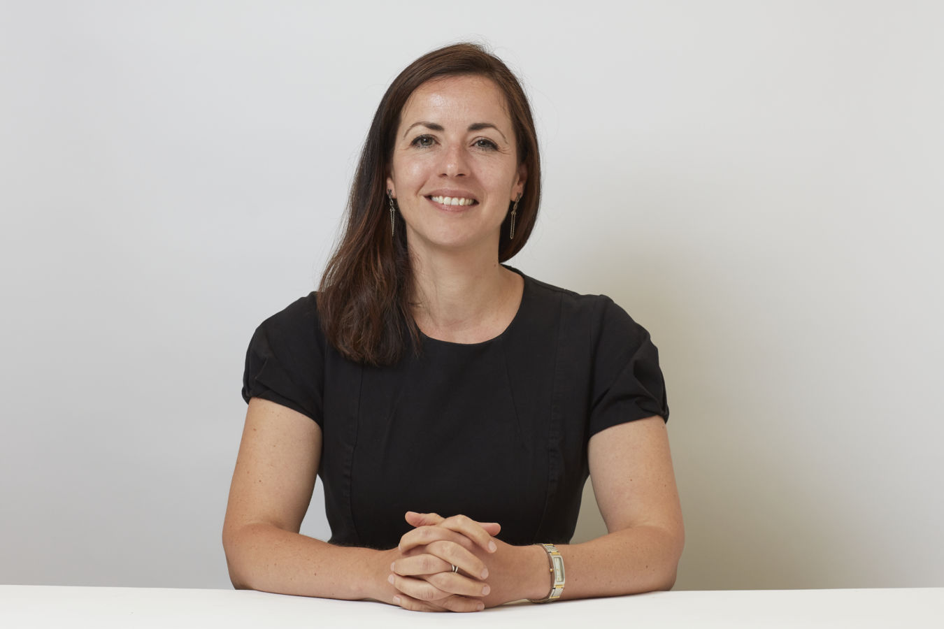 Woman sitting at a white table with hands clasped, wearing a black shirt and a watch, smiling against a plain white background.