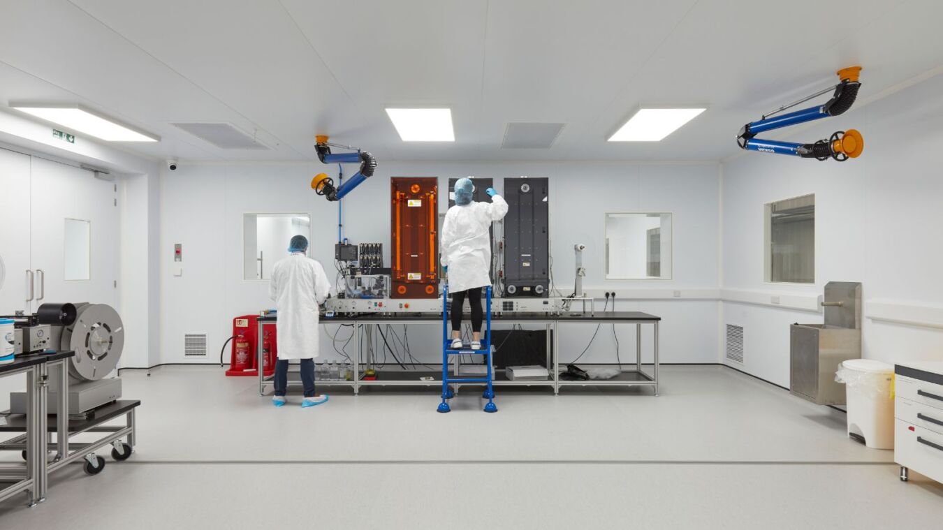 Two technicians in cleanroom attire operate machinery in a bright, sterile laboratory with overhead ventilation and various equipment on tables and carts. Red warning signs and fire extinguishers are visible.