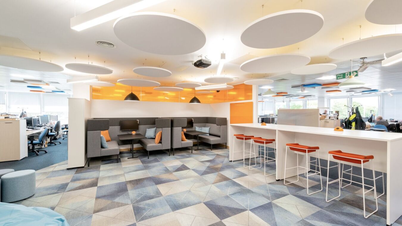 Vibrant office lounge with booth seating, high tables and stools, circular ceiling panels, geometric floor tiles, and orange accent lighting.
