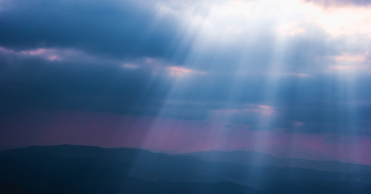 Sunbeams shine through thick clouds, illuminating the shadowy mountain range below, creating a serene, ethereal atmosphere with a pink and blue-tinted sky.