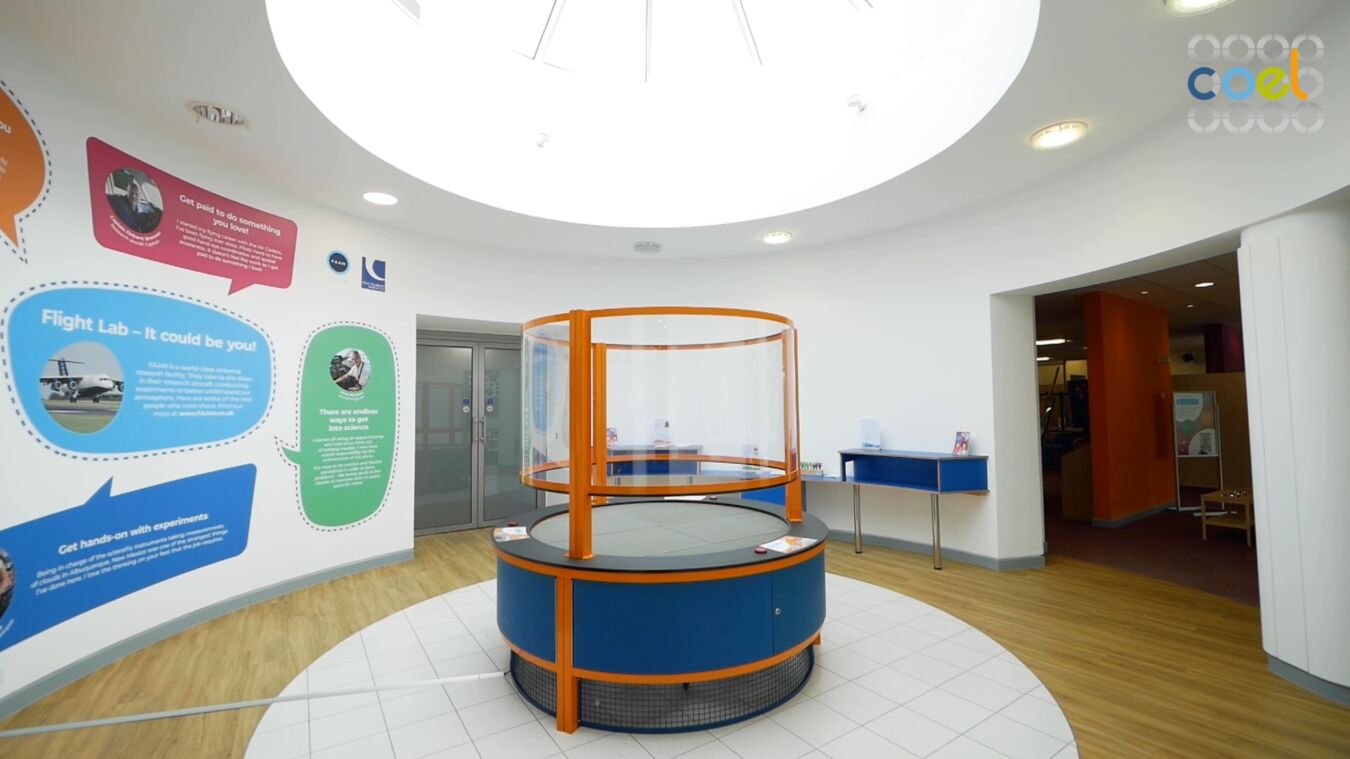 Circular display table with orange frame and clear panels stands unused in a brightly lit lobby. Surrounding walls feature educational text: “Flight Lab – It could be you!” Various educational posters and brochures are visible.