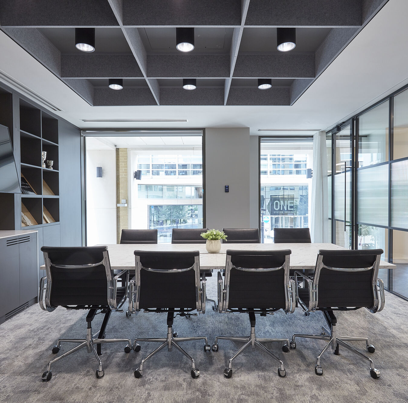 Modern conference room with a long white table, eight black office chairs, floor-to-ceiling windows, and geometric ceiling panels with recessed lighting.