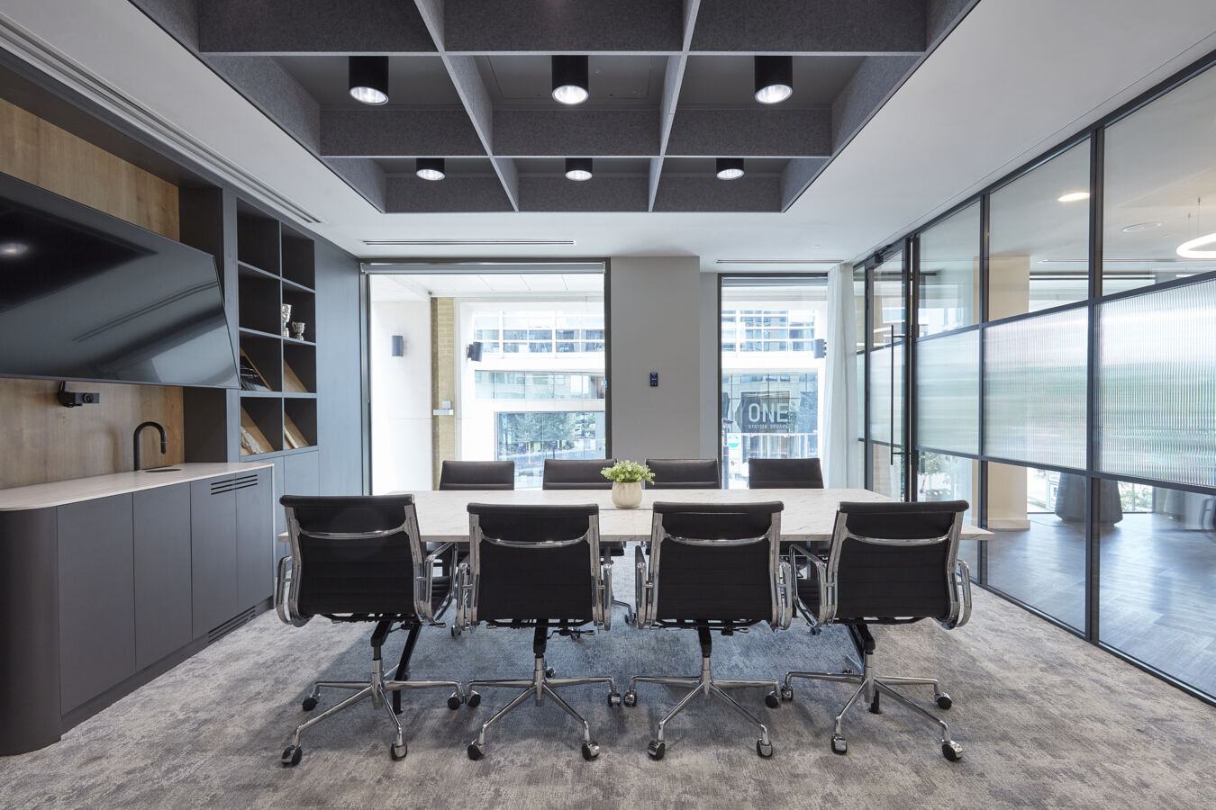 Modern conference room with a long white table, eight black office chairs, floor-to-ceiling windows, and geometric ceiling panels with recessed lighting.
