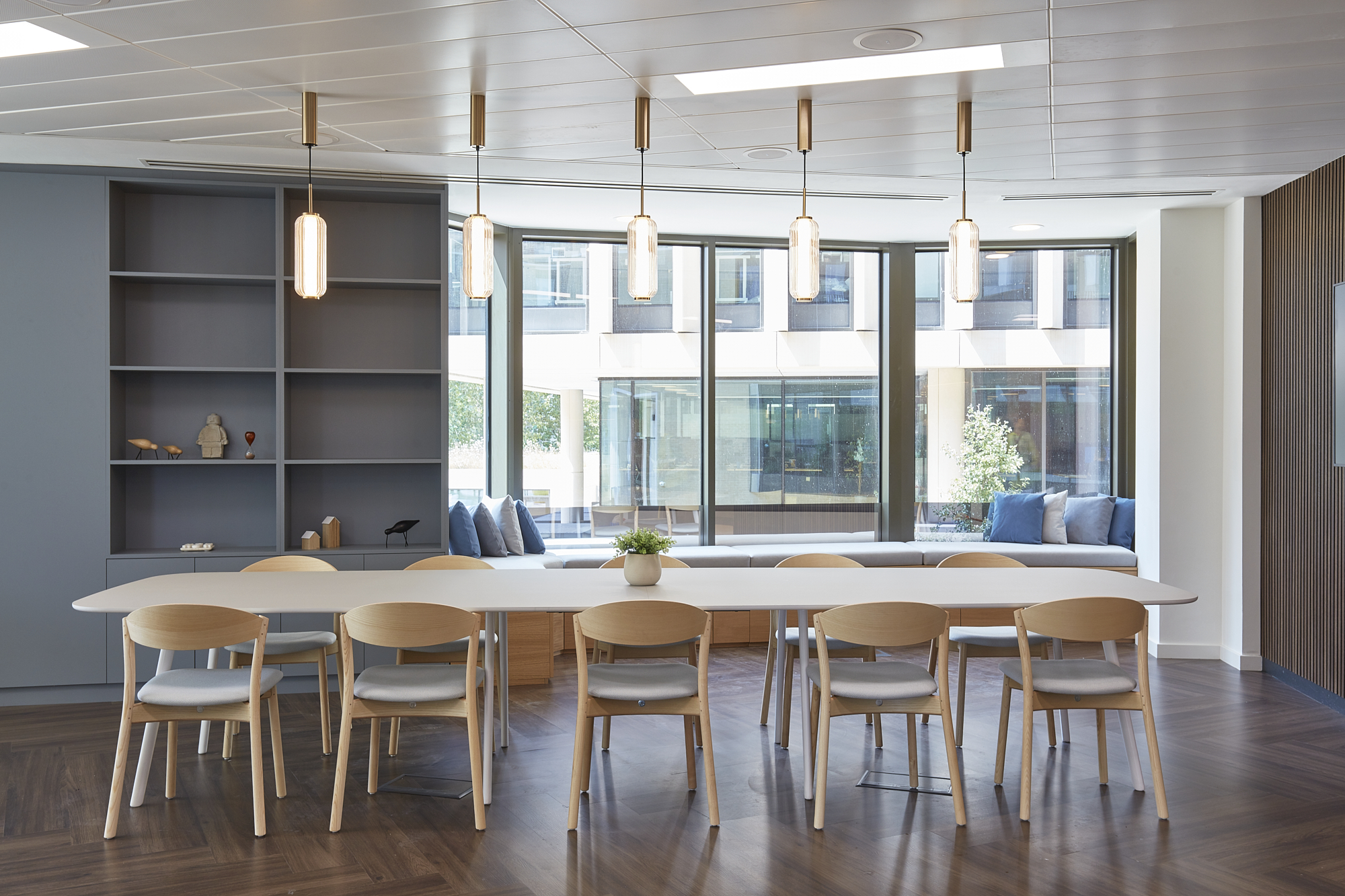 A long white table with wooden chairs is centrally placed under pendant lighting in a modern room, featuring shelves, large windows, and cushioned seating with blue and gray pillows.