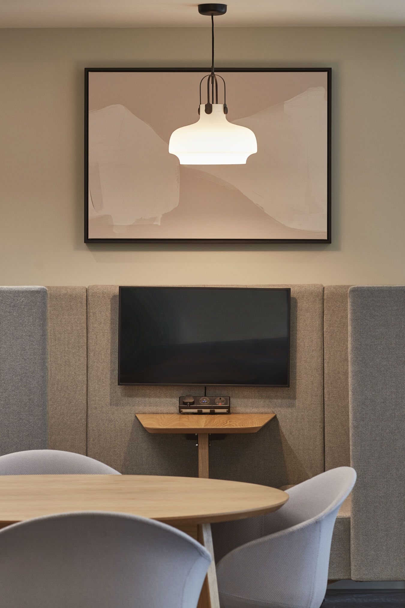 Modern meeting space with a round table and four chairs, a small wall-mounted TV between acoustic panels, and a pendant light hanging above. A framed abstract artwork is displayed on the wall.