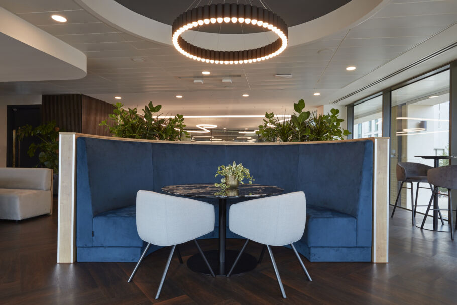 Stylish booth seating with a round table, two white chairs, overhead ring light fixture, and greenery in the background.