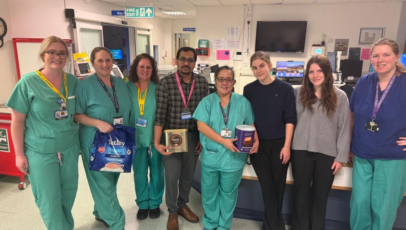 Group of healthcare workers standing together, some in scrubs, holding containers of Tetley tea, Nescafé coffee, and Cadbury hot chocolate, inside a hospital ward with medical equipment.