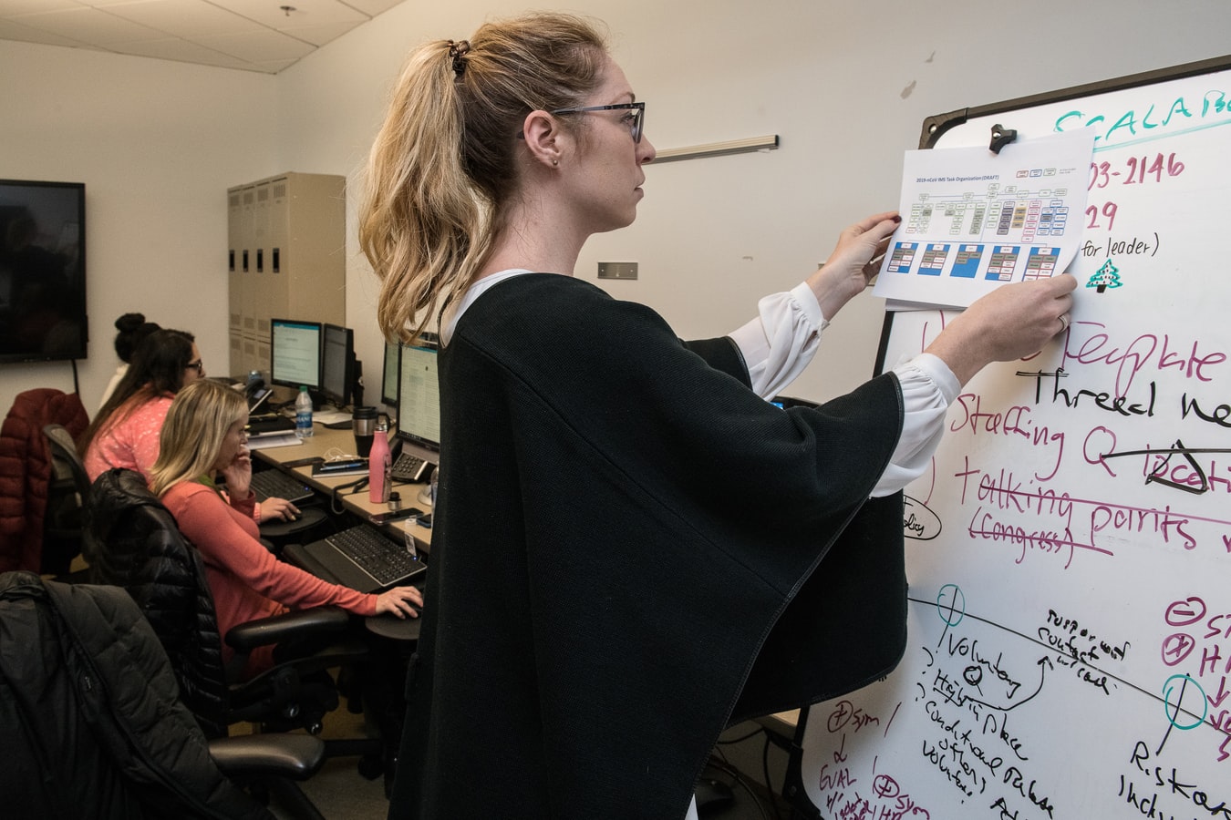 A woman presents a chart on a whiteboard covered in colorful writing, while others work at computers in an office setting.
