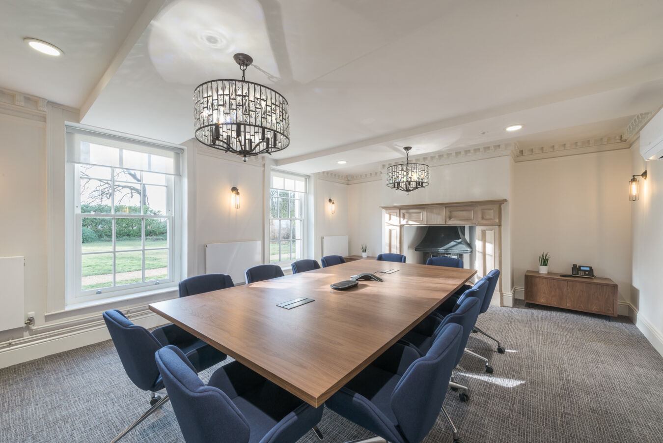 Conference room featuring a large wooden table surrounded by blue chairs; elegant ceiling lights hang above, and large windows provide natural light, overlooking a grassy landscape.