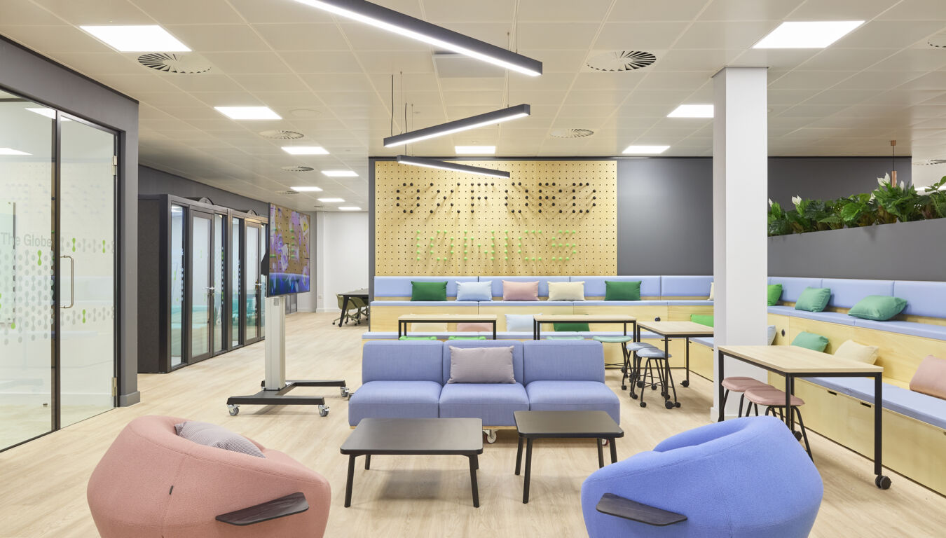 Modern office lounge with pastel furniture, including blue sofas and pink beanbag chairs, arranged around tables. Pegboard wall with plants and text "The Globe" visible through glass door.