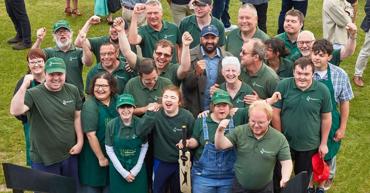 A group wearing matching green Phoenix Bites shirts and aprons, raising fists in celebration, stands on green grass. They appear joyous and united, likely at an outdoor event.