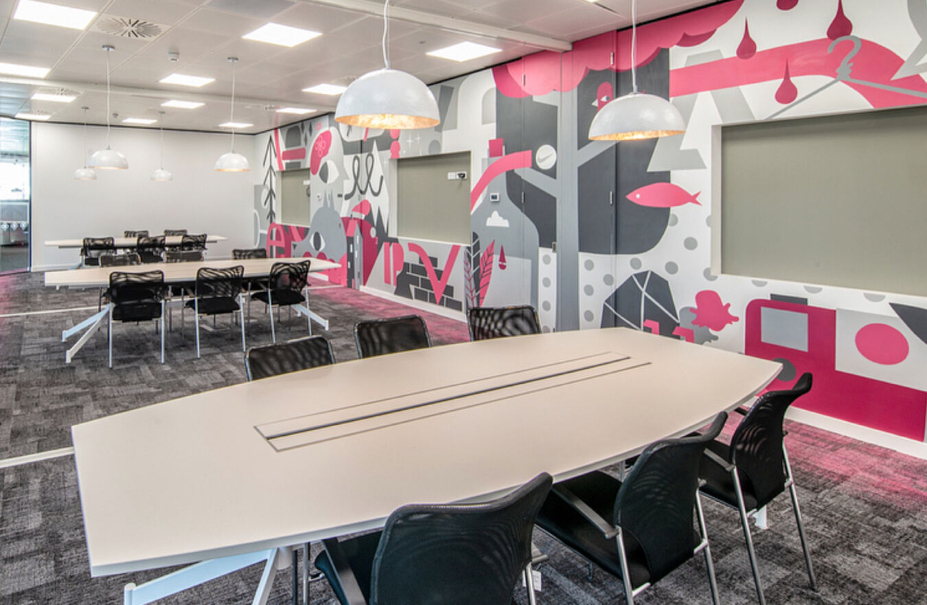 Modern conference room with sleek tables and black chairs, featuring a colorful abstract mural and pendant lighting, creating a vibrant, contemporary atmosphere.