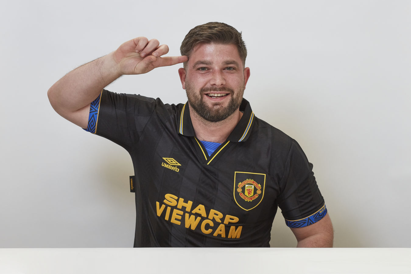 A man wearing a black Umbro football jersey, with "SHARP VIEWCAM" text and a Manchester United logo, smiles and salutes, seated against a plain white background.