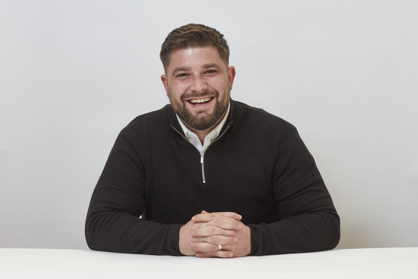 A man wearing a black sweater sits smiling with hands clasped on a white table against a plain white background.