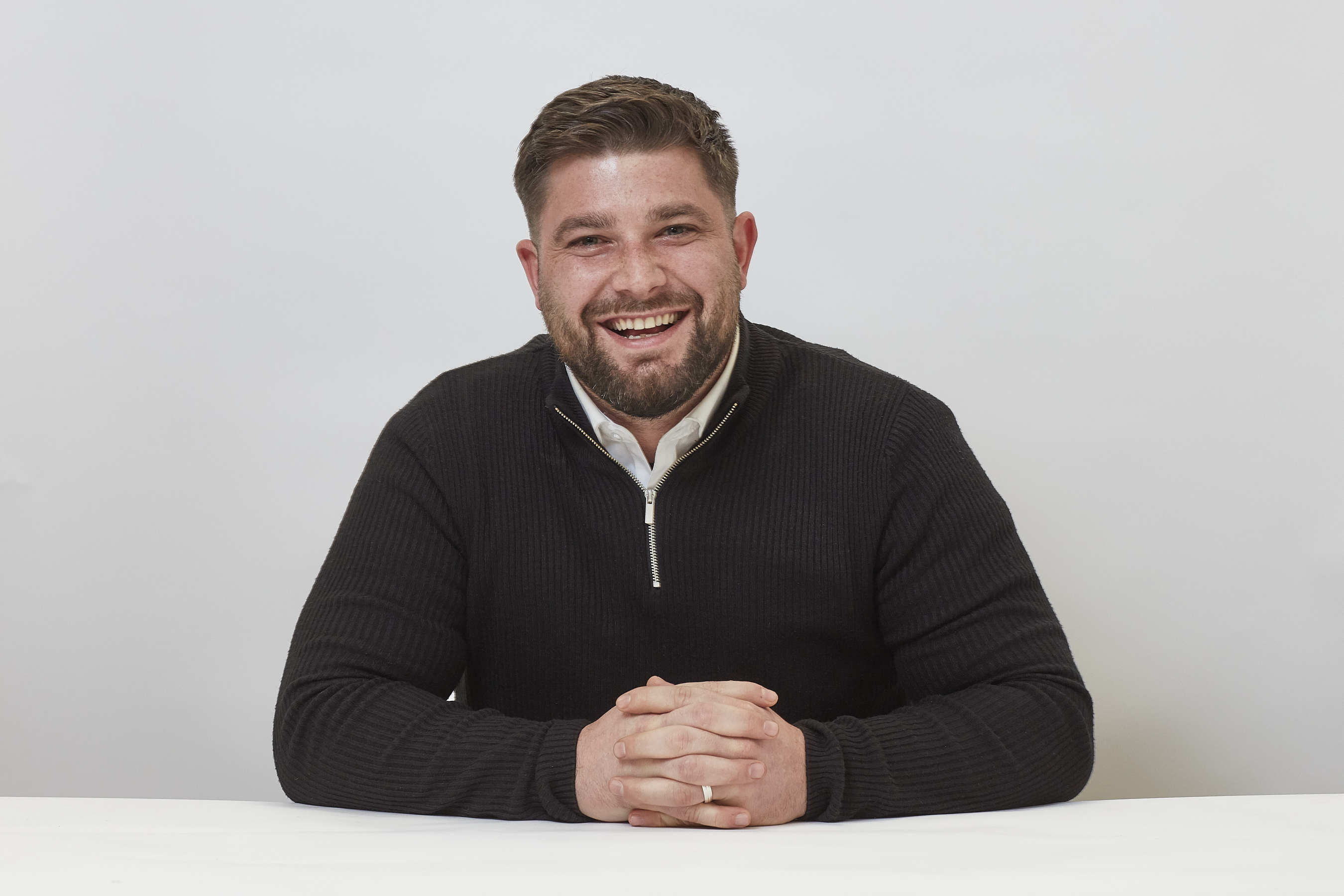 A man wearing a black sweater sits smiling with hands clasped on a white table against a plain white background.