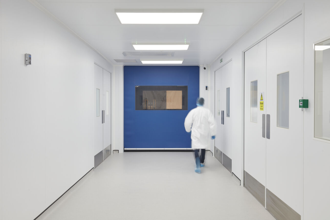 Person in a white lab coat walks down a sterile, brightly lit laboratory corridor with white walls, blue accents, and a sign on the door reading "Danger Laser in Use" in English.