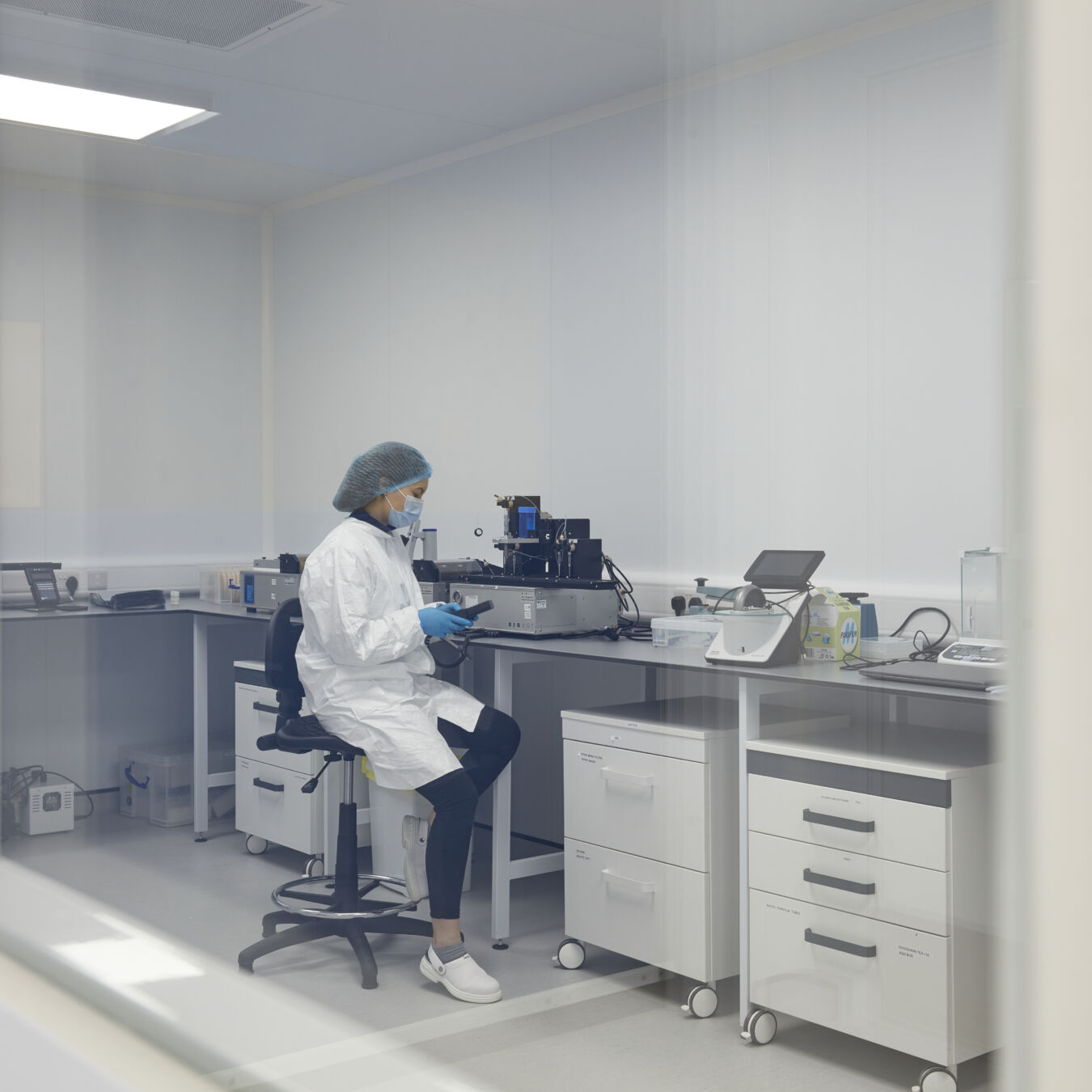 A lab technician, wearing protective attire and gloves, works with a device on a counter in a sterile lab environment with white cabinets and equipment.