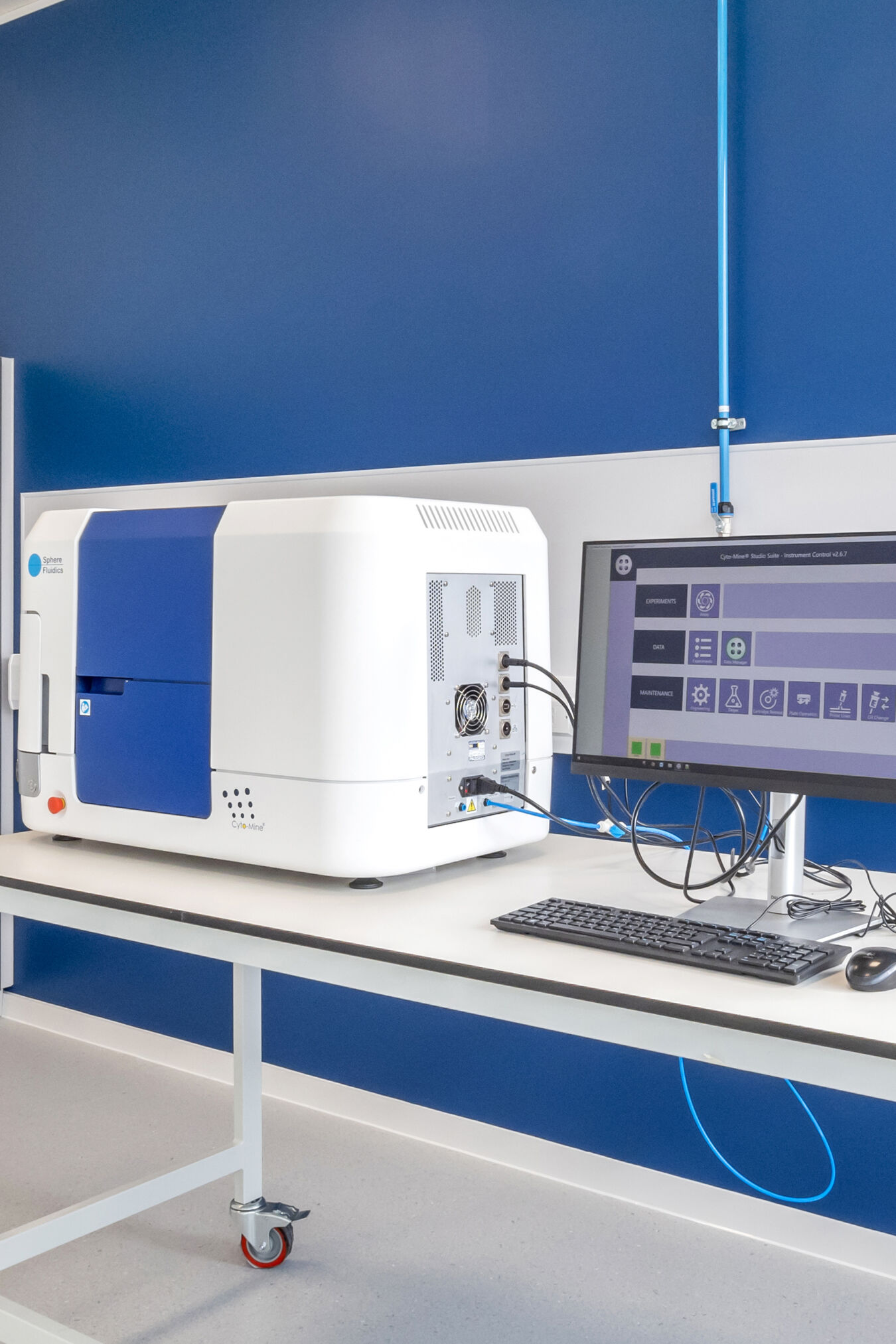 A laboratory instrument sits on a table, connected to a monitor displaying a control interface, with blue and white walls and a gray cabinet in the background.