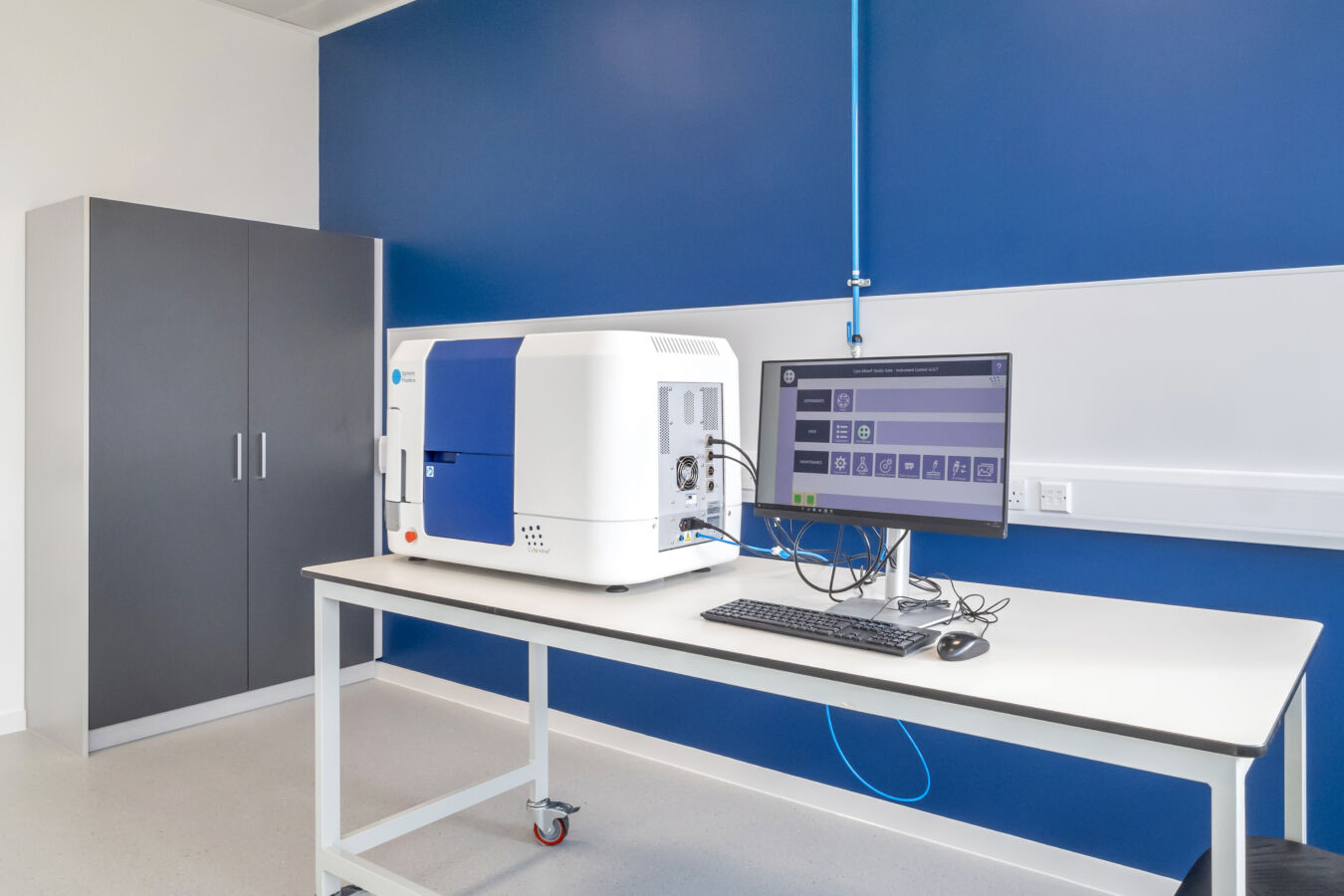A laboratory instrument sits on a table, connected to a monitor displaying a control interface, with blue and white walls and a gray cabinet in the background.