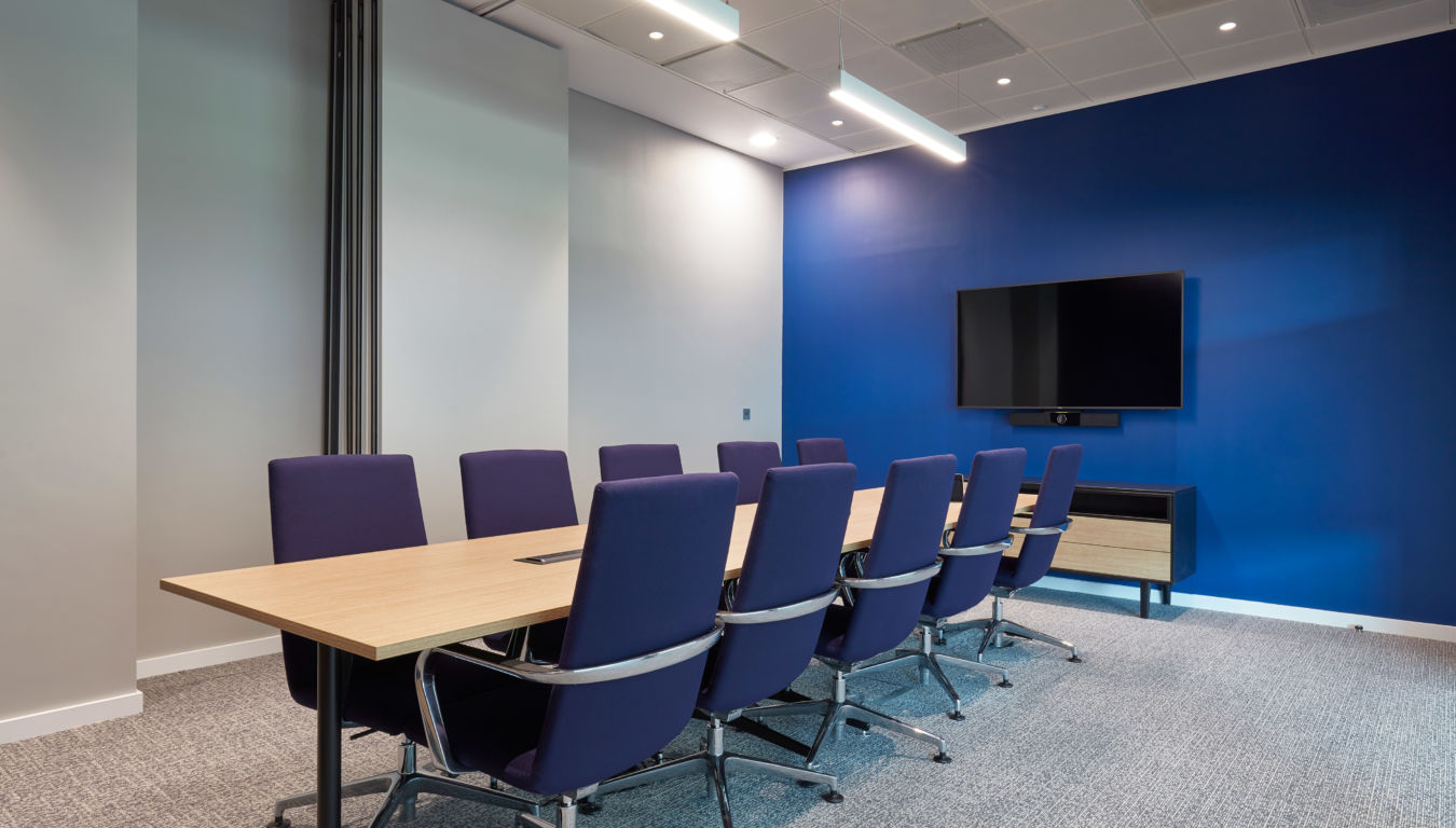 Modern conference room with a long wooden table, eight blue chairs, and a flat-screen TV on a blue accent wall. Bright lighting and gray carpet.