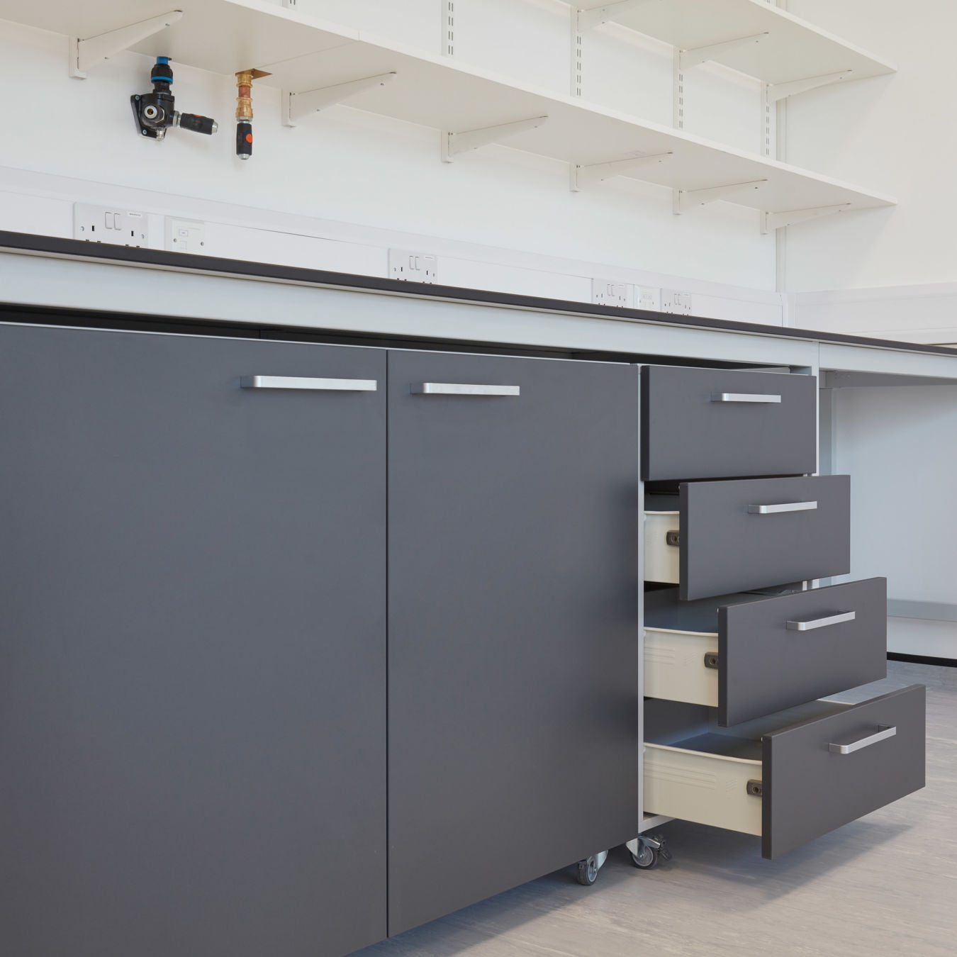 Close-up view of laboratory cabinetry with dark grey drawers and cupboards, some of which are open, beneath a white work surface and wall-mounted shelves